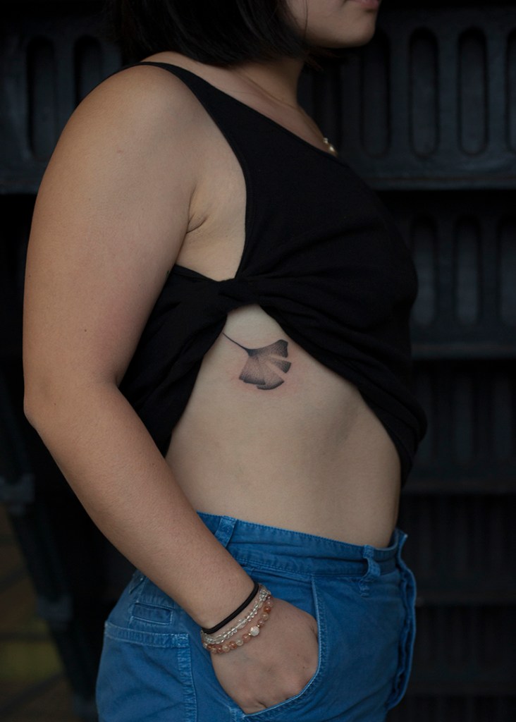 handpoke tattoo van een ginkgo blad rechts op de torso, vlak onder de bh band. Het blad ziet er breekbaar uit met veel gradaties