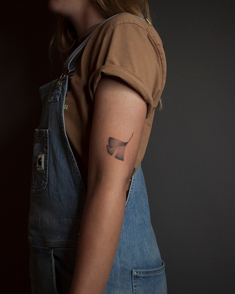 handpoke tattoo van een ginkgo blad op de buitenkant van het linker bovenarm, net boven de elleboogsplooi. Het blad ziet er breekbaar uit met veel gradaties