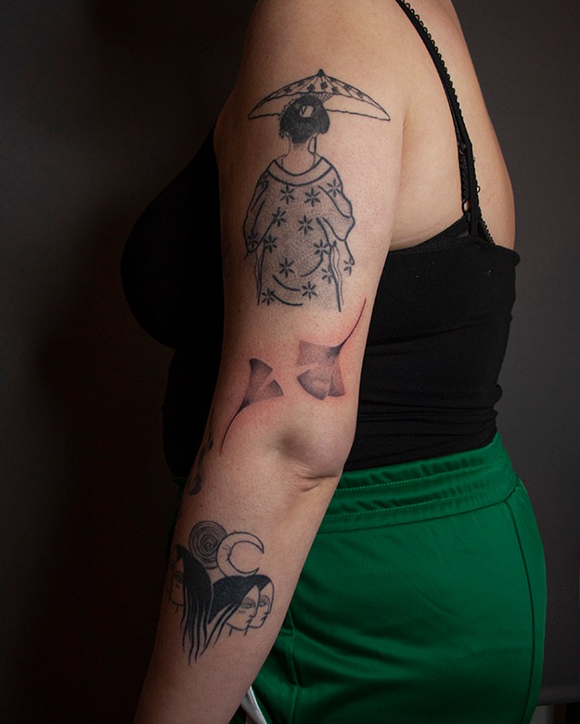 handpoke tattoo van twee ginkgo bladeren op de buitenkant de linker bovenarm, vlak boven de elleboogsplooi. De bladeren zien er breekbaar uit met veel gradaties