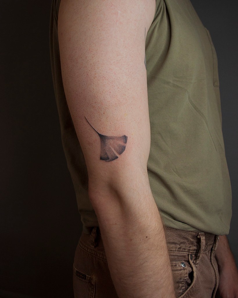 handpoke tattoo van een ginkgo blad op de buitenkant de rechter bovenarm, net boven de elleboogsplooi. Het blad ziet er breekbaar uit met veel gradaties