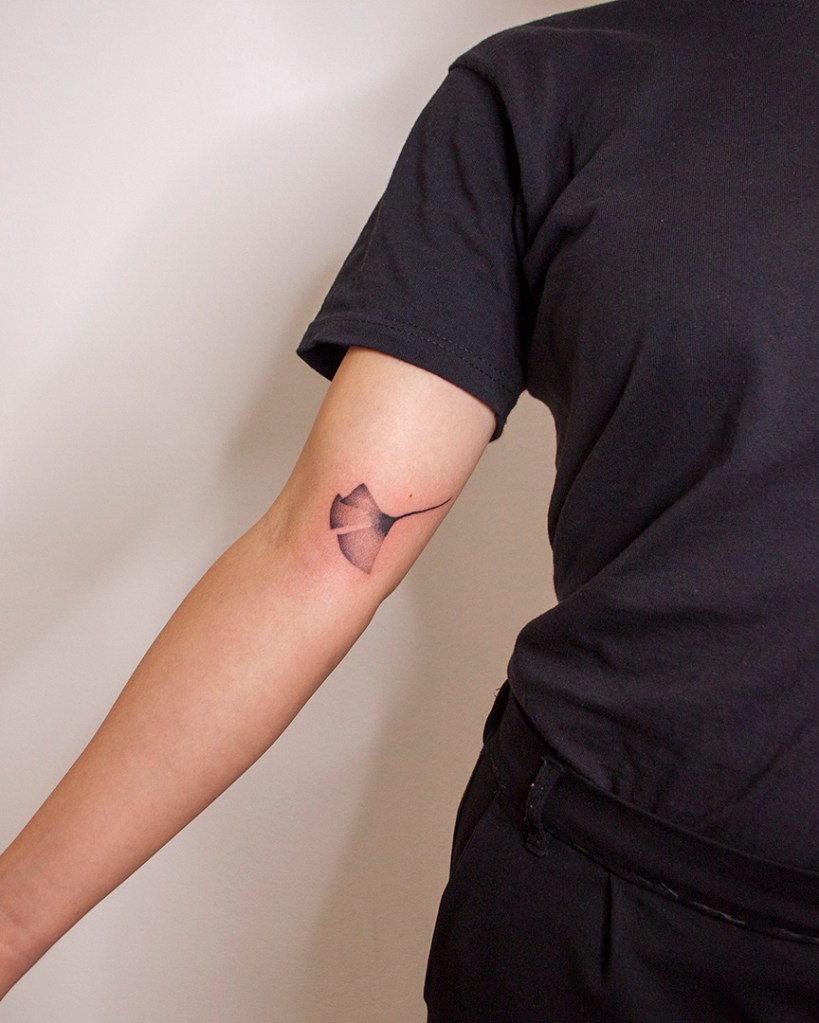 handpoke tattoo van een ginkgo blad op de binnenkant van de rechter bovenarm, tussen de bicep en de elleboogsplooi. Het blad ziet er breekbaar uit met veel gradaties