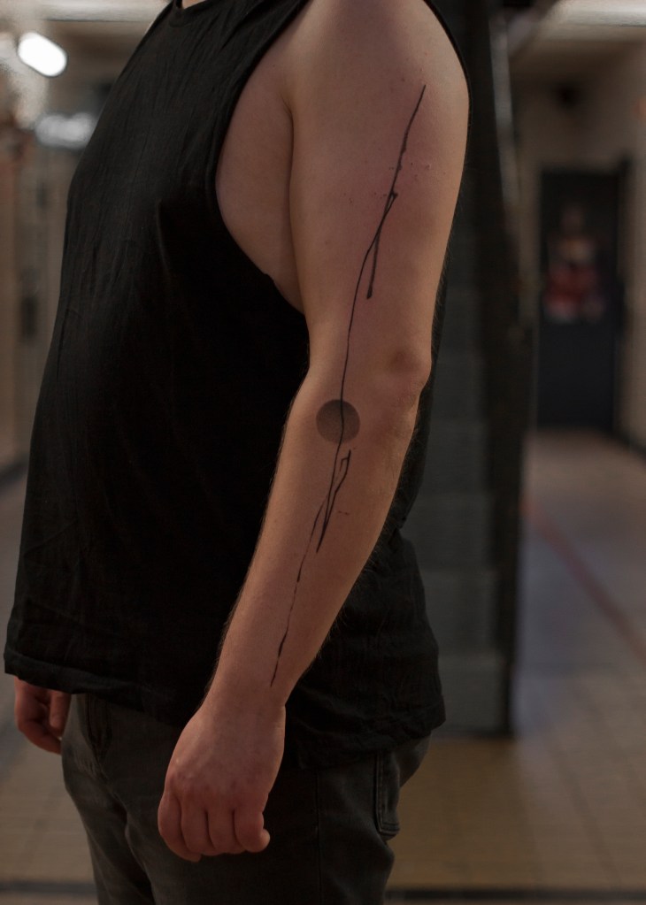 Tattoo op de buitenkant van de linkerarm. Er is een lijn te zien er asl inkt op lijkt geschilderd, er is een cirkel vorm op de achtergrond